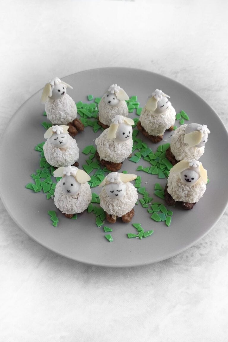 Plate with nine coconut-coated sheep-shaped desserts, each with almond ears and chocolate faces, surrounded by green chocolate shard decorations.