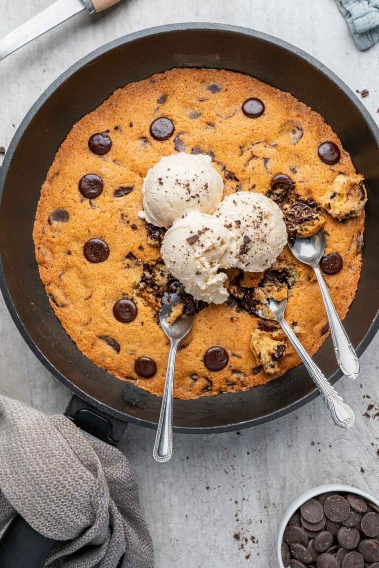 Overhead shot of a warm chocolate chip skillet cookie in a black skillet, topped with scoops of vanilla ice cream and three spoons.