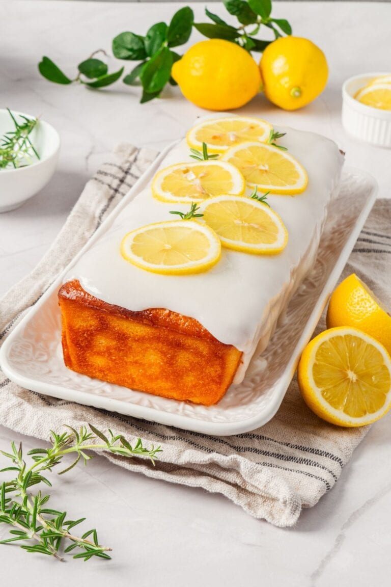 A glazed lemon loaf cake garnished with lemon slices and rosemary on a white plate.