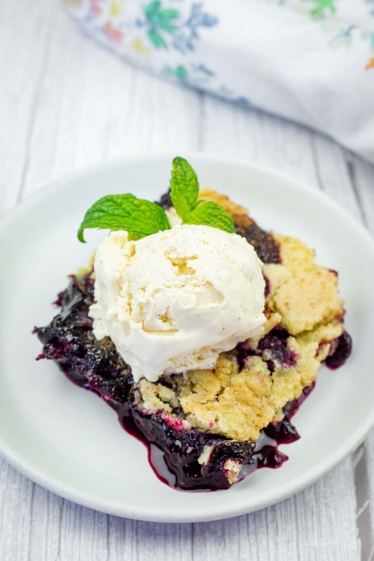 A white plate holds a serving of blueberry dump cake topped with a scoop of vanilla ice cream and fresh mint.