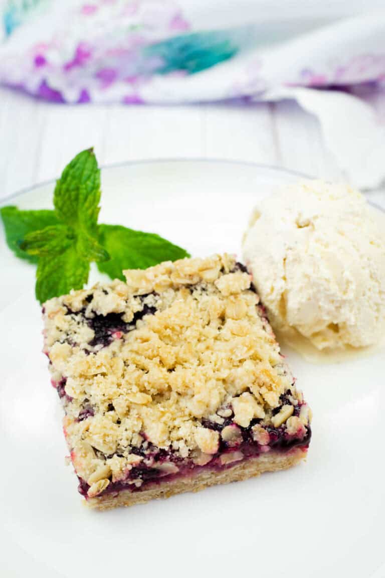 A white plate holds a square Blueberry-Rhubarb Crumble Bar topped with a scoop of vanilla ice cream and fresh mint, with a colorful cloth in the background.