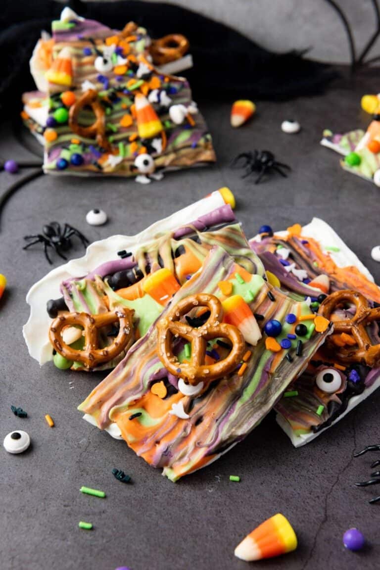 Colorful Halloween-themed bark topped with pretzels, candy eyes, candy corn, and sprinkles, displayed on a dark surface with plastic spiders and web decorations.