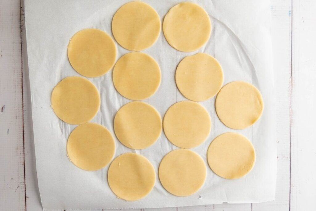 Fourteen round pieces of rolled dough are arranged on a sheet of parchment paper.