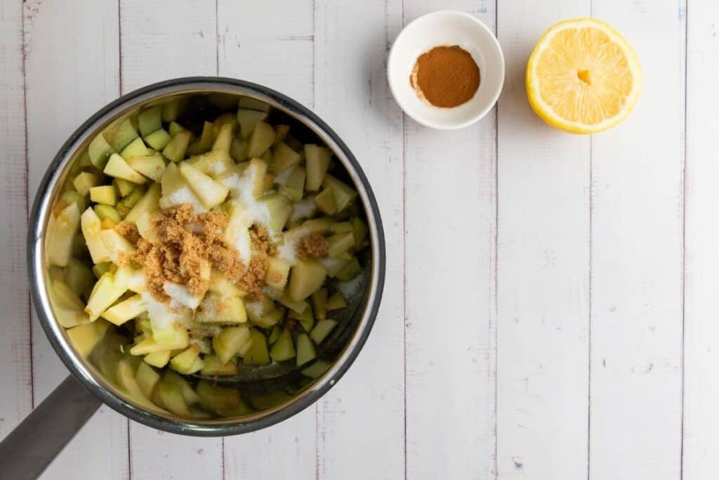 A saucepan with chopped apples, white and brown sugar. Nearby are a bowl of cinnamon and a halved lemon on a white wooden surface.