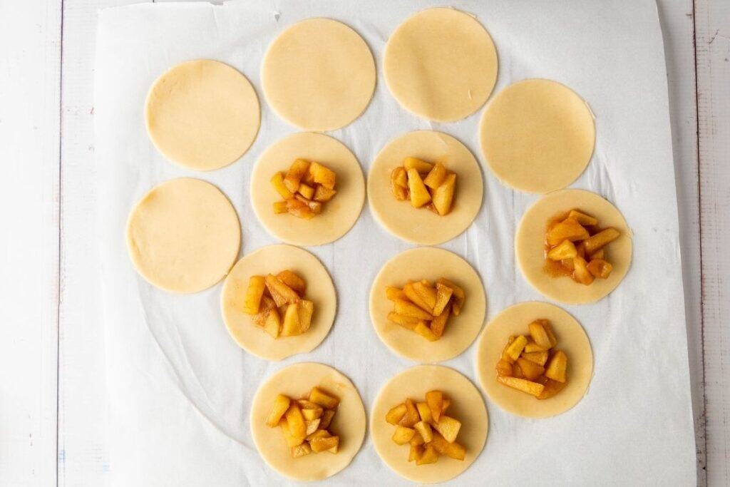 Twelve dough circles on parchment paper, six topped with a filling of chopped fruit, likely apples, and six left plain.