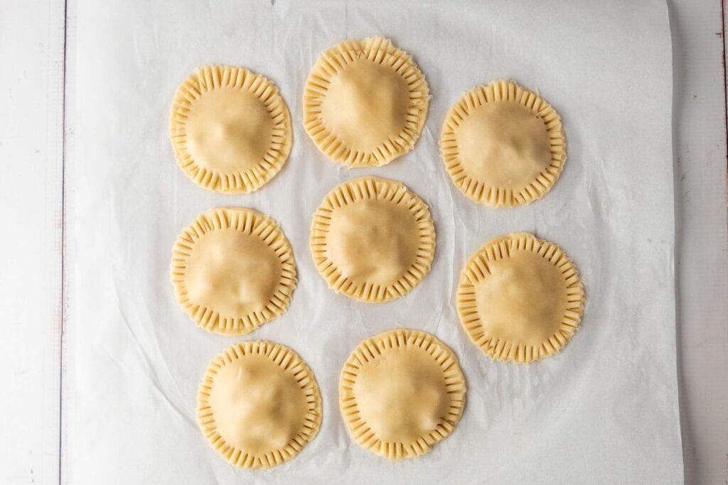 Eight uncooked round hand pies with crimped edges are arranged in neat rows on a sheet of parchment paper.
