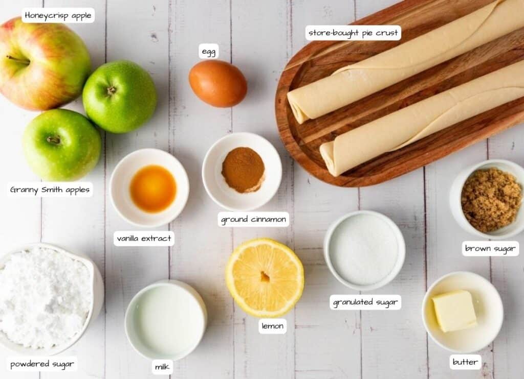 Ingredients for apple pie arranged on a white surface, including apples, pie crust, egg, vanilla, cinnamon, lemon, sugars, butter, and milk, each labeled with text.