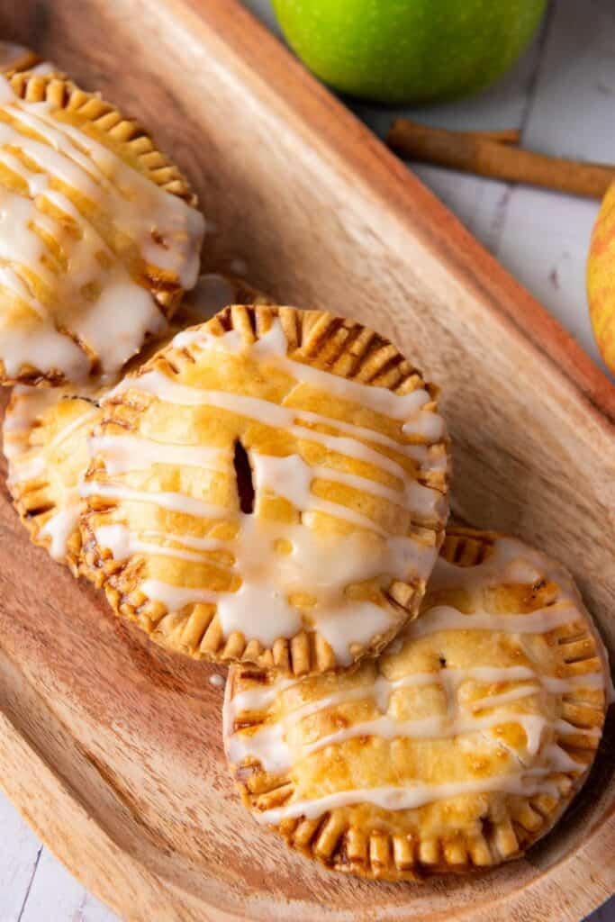 Three small round pastries with icing drizzle are arranged on a wooden tray next to an apple.
