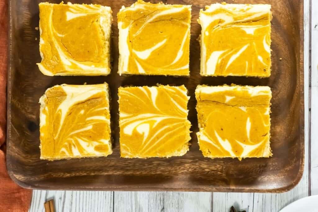 Six pieces of Pumpkin Cheesecake Bars on a wooden plate placed on a white wooden table with napkin.