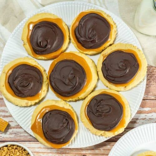 Seven pieces of Crumbl Copycat Twix Cookies on a white plate on a wooden table.