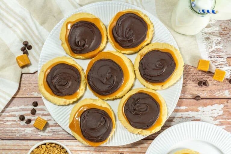 Seven pieces of Crumbl Copycat Twix Cookies on a white plate on a wooden table.