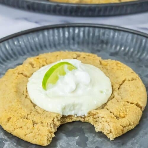 A large Copycat Crumbl Key Lime Cookies with a bite taken out, topped with white cream and a lime wedge, served on a gray plate.
