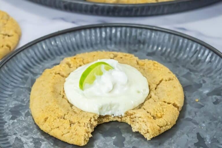A large Copycat Crumbl Key Lime Cookies with a bite taken out, topped with white cream and a lime wedge, served on a gray plate.