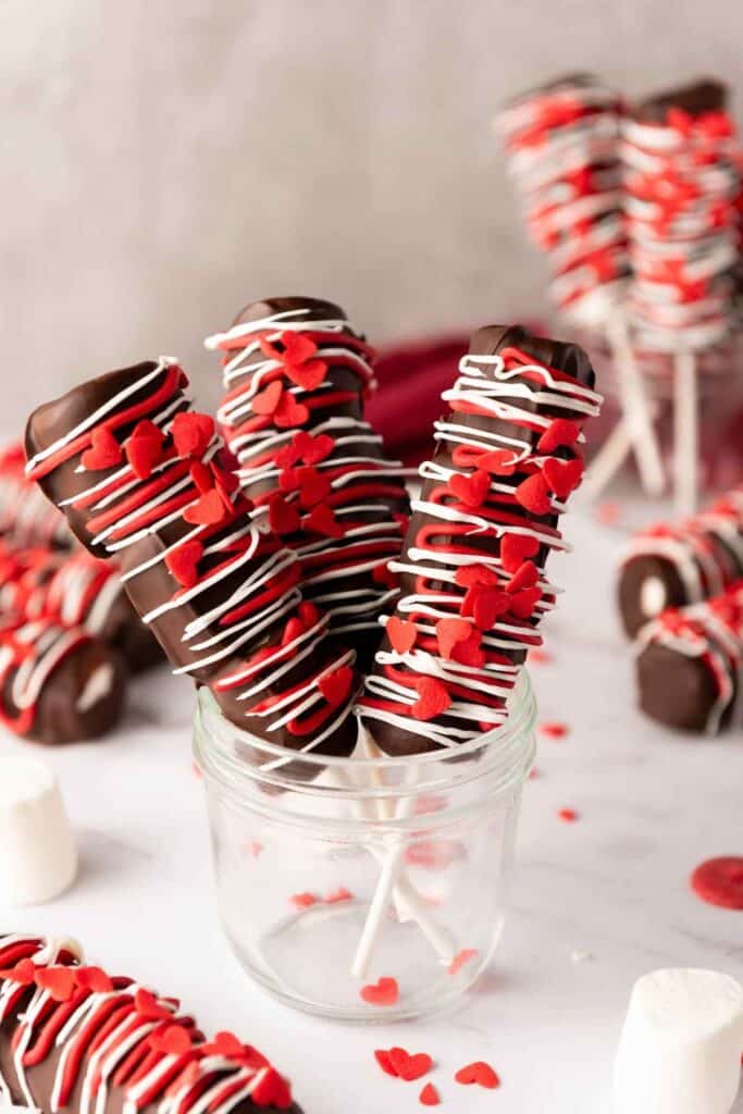 Decorated marshmallow pops with chocolate, red and white icing, and heart-shaped sprinkles, perfect for Valentine's Day.